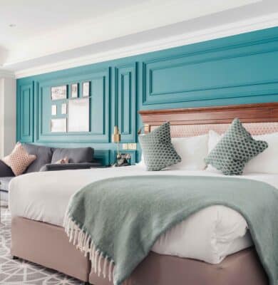 Elegant hotel room with plush king bed, stylish teal decor, cosy seating, window views, and modern amenities.