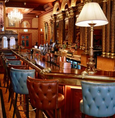 Elegant hotel bar with luxurious tufted chairs, ornate mirrors, warm lighting, and a sparkling chandelier.