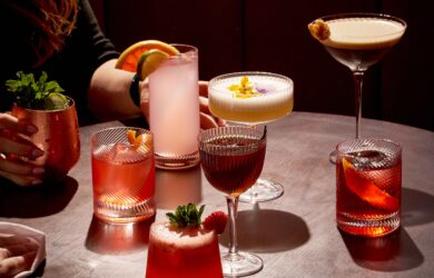 Guests enjoy vibrant cocktails on a stylish wooden table in a chic bar with plush red seating, perfect for relaxing evenings.