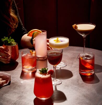 Guests enjoy vibrant cocktails on a stylish wooden table in a chic bar with plush red seating, perfect for relaxing evenings.