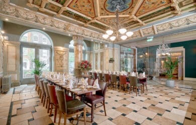 Elegant hotel dining room with ornate ceiling, large windows, and set tables, offering a refined dining experience.
