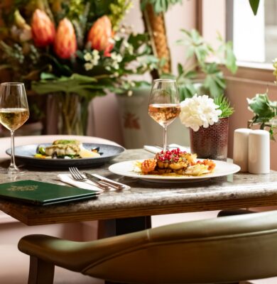 Elegant dining table with gourmet dishes, wine glasses, vibrant flowers, and lush greenery in a cosy hotel restaurant.